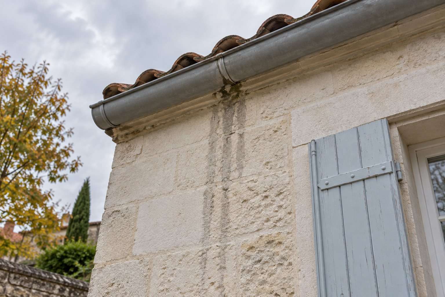 Coulures sur un mur clair sous le raccord d’une gouttière métallique, à proximité d’un volet bleu pâle.